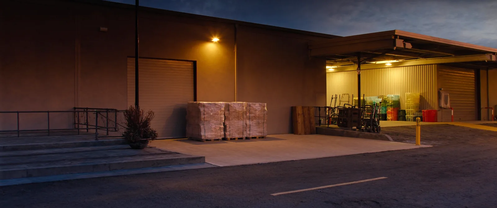 The shipping dock at dusk, three pallets banded and waiting under the light, forklifts parked back under the canopy.