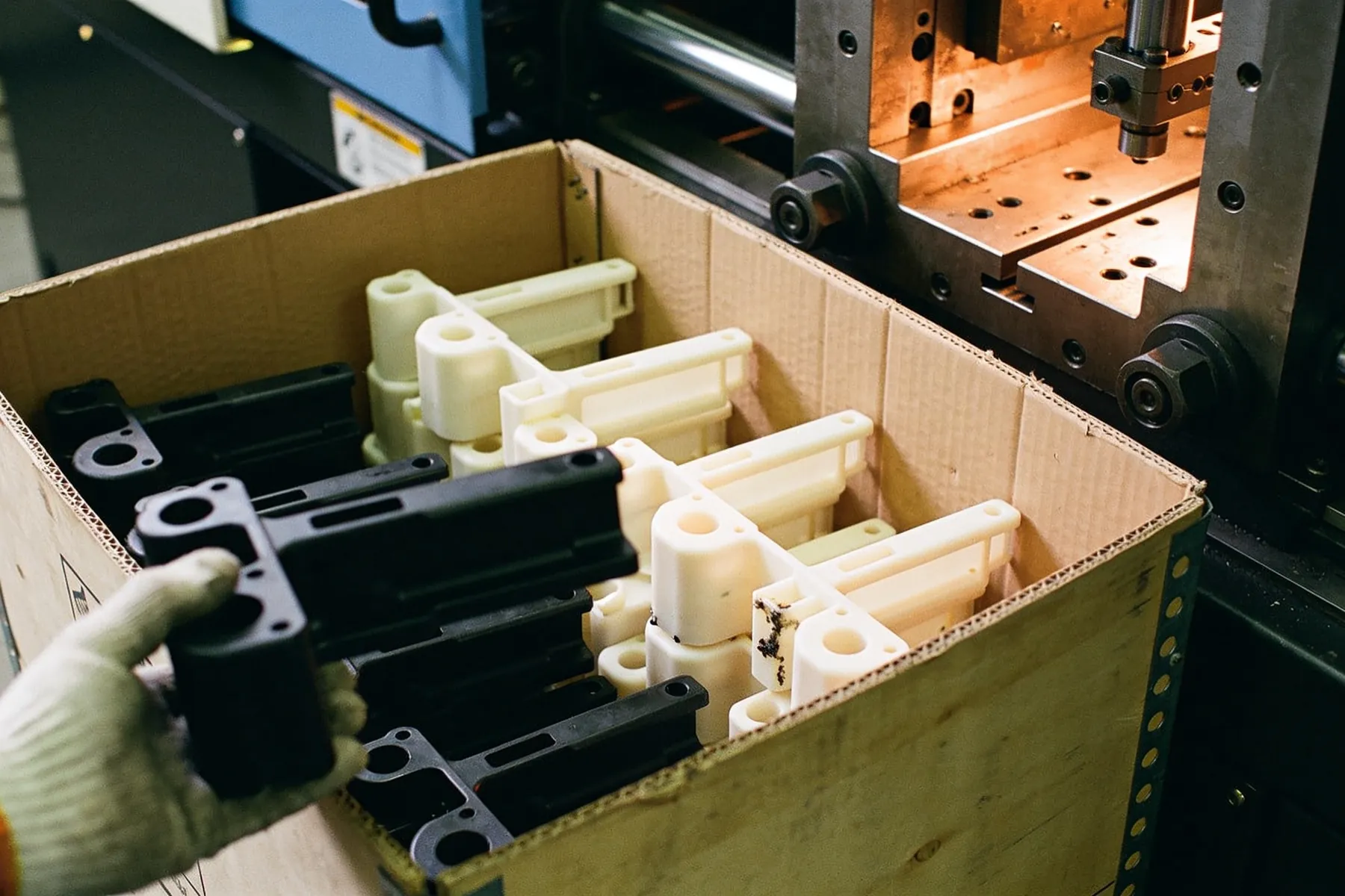 A gloved hand packing black and natural moulded brackets upright into a divided corrugated box beside the press.